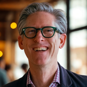 Smiling middle-aged person with glasses, wearing a blazer, in a softly lit indoor setting.